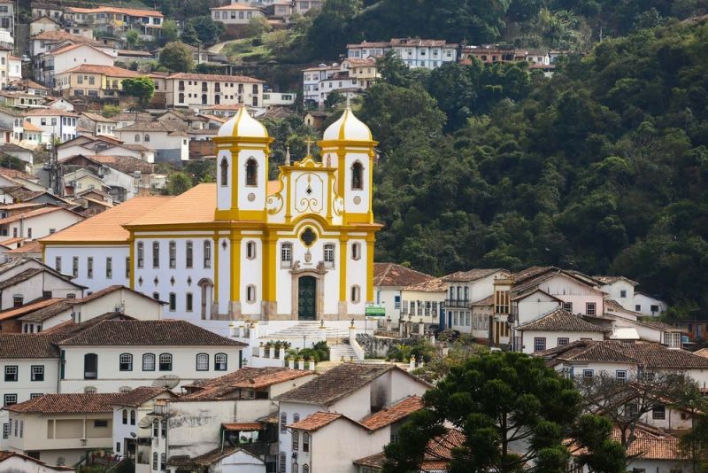 Igreja histórica em Ouro Preto com fachada branca e detalhes dourados, rodeada por casas coloniais e vegetação de serra